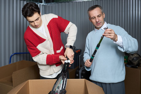 Caucasian teenager man and middle aged Caucasian man sorting tennis rackets together, standing near...