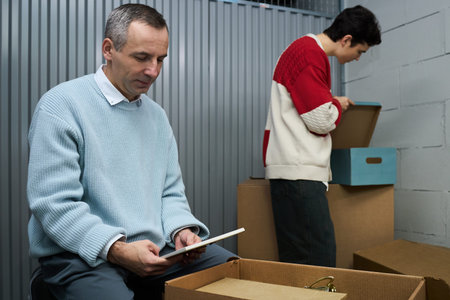 Middle aged Caucasian man sitting and looking at photograph while young adult man with dark hair, standing and unpacking cardboard box in storage unit with corrugated metal wallの写真素材