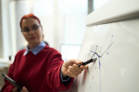 Caucasian young adult woman explaining graph on whiteboard, holding pen and gesturing toward data...