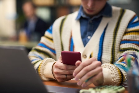 Caucasian teenager boy using smartphone while sitting at desk with open laptop, focusing on mobile...