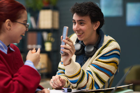 Young man smiling while showing smartphone screen to young woman, both sitting at table in casual...