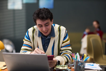 Young adult Caucasian man sitting at desk smiling while using smartphone and working on laptop in...
