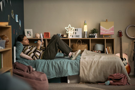 Teenage Caucasian boy lying on bed holding smartphone, listening to tutor during online lesson,...