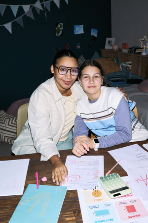Vertical of Caucasian teenage girl sitting beside Black woman tutor, both smiling and looking at...