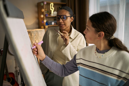 Young Black woman tutor observing and guiding teenage Caucasian girl writing on whiteboard during...