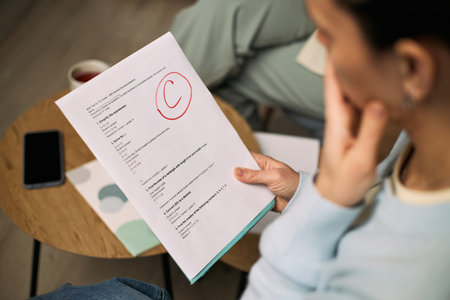 Caucasian young adult woman holding test paper with large red C grade, sitting at table during...