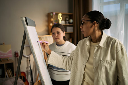 Black woman tutor explaining concept on flip chart while teenage Caucasian girl student attentively...