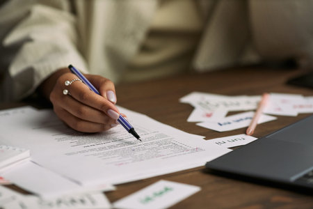 Caucasian young adult woman holding pen marking printed lesson materials on desk near laptop,...