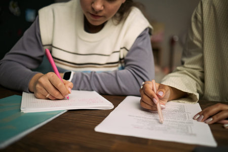 Teenage girl and woman studying together at table, writing in notebooks and reviewing printed...