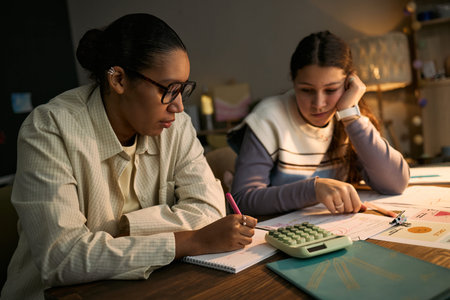 Young adult Black woman tutoring teenage Caucasian girl, both sitting at desk working on math...