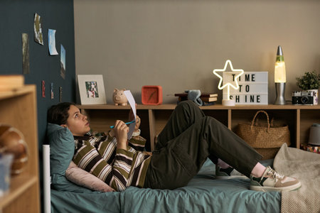 Teenage girl lying on bed studying lesson notes, holding pen and paper, looking thoughtful,...
