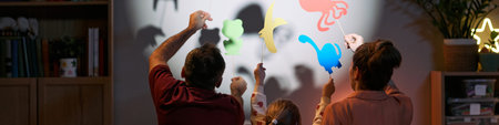 Caucasian man, Caucasian woman, and child creating animal shadow puppets on wall, raising colorful paper cutouts together, backs facing camera, engaging in playful family activityの写真素材