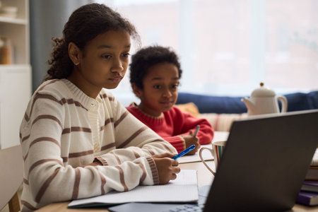 Black preteen girl sitting at table using laptop and writing in notebook while Black younger boy sitting beside her watching screen, both focused on online learning or homeworkの写真素材