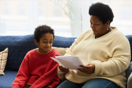 Black senior woman sitting on sofa showing documents to Black child, both engaging in conversation, child looking at papers with interest, home environment visible in backgroundの写真素材