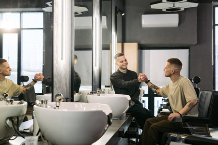 Two young adult Caucasian men greeting each other with handshake in modern barbershop, one sitting...