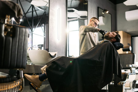 Caucasian young adult man reclining in barber chair receiving beard trim from Caucasian young adult...
