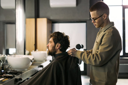 Caucasian young adult man cutting hair of Caucasian middle aged man in modern barbershop, barber...