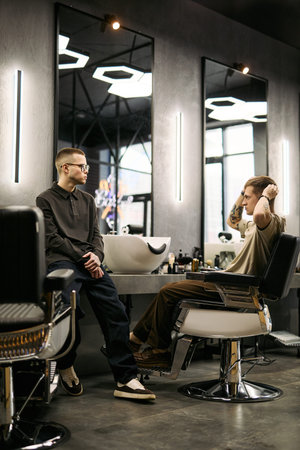 Young Caucasian man sitting in barber chair adjusting hair while young Caucasian male barber, standing nearby watching client in modern barbershop with mirrors and salon equipment visibleの写真素材