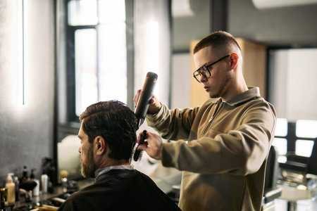 Young adult man standing styling hair of middle aged man using hair straightener and comb in modern...