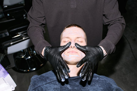 Caucasian young adult man receiving facial treatment from male barber wearing black gloves in...