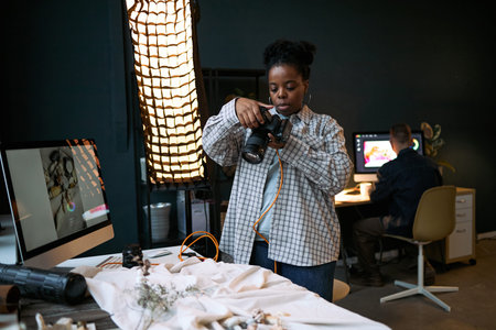 Black woman young adult adjusting camera settings while standing at creative workspace with computer...