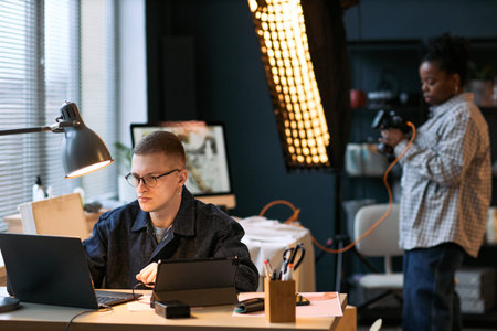 Caucasian young adult man working on laptop at desk, Black young adult woman standing in background...