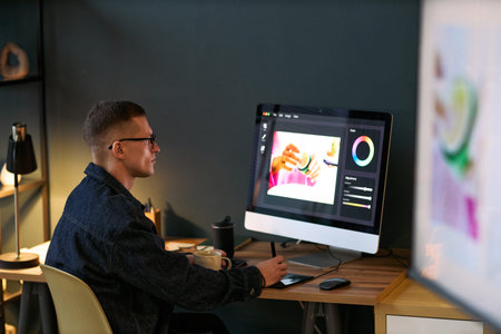 Young adult man working at desk using graphic tablet and large computer monitor, editing digital...