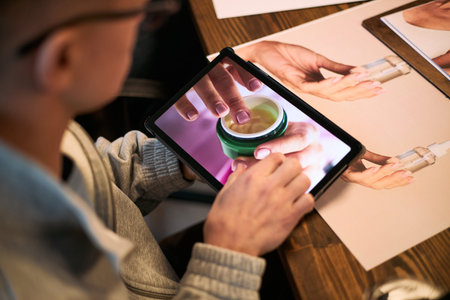 Young adult man using tablet, viewing close up image of hands applying skincare cream, sitting at...