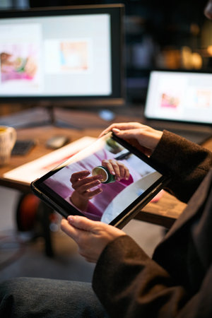 Woman holding digital tablet displaying close up of hands holding skincare product, sitting at desk...