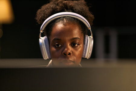 Portrait of young adult Black woman wearing headphones concentrating while looking at computer...