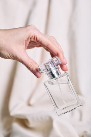 Caucasian female hand holding transparent perfume bottle against neutral background, fingers gently...