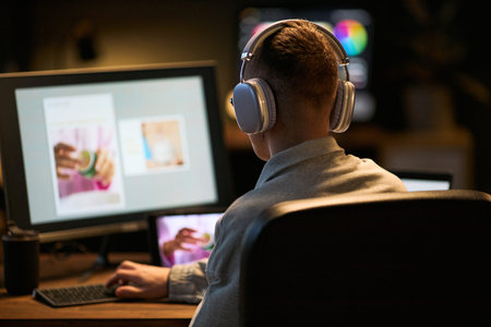 Young adult man wearing headphones sitting at desk working on computer with large monitor displaying...