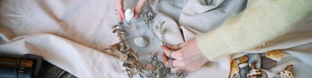 Caucasian young adult woman arranging dried plants and decorative objects on fabric surface, hands visible holding glass dropper bottle and branch, natural elements scattered nearbyの写真素材