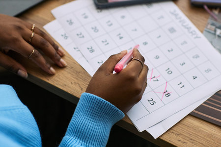 Black woman marking dates on calendar with pink pen, focusing on specific days in January,...