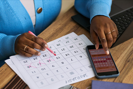 Young adult Black woman marking menstrual cycle dates on paper calendar while using period tracking app on smartphone, sitting at desk with laptop, focusing on health managementの写真素材