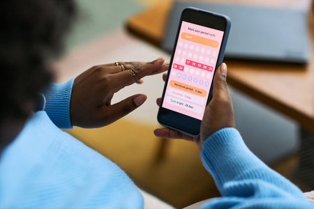 Black woman using smartphone tracking menstrual cycle with femtech health app, hand interacting with period calendar interface, focusing on reproductive health managementの写真素材
