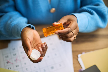Black woman opening prescription pill bottle, holding medication in hand, sitting at desk with...