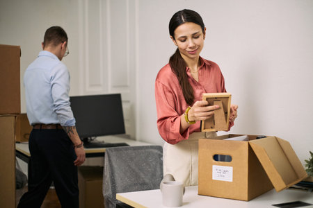 Young adult Caucasian woman unpacking framed photo while cleaning office workspace, middle aged...