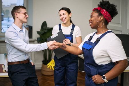 Caucasian young adult man shaking hands with Black young adult woman in cleaning uniform, while Asian young adult woman in cleaning uniform holding clipboard and smiling in office settingの写真素材