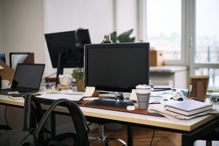 Empty office workspace showing computer monitor, keyboard, reusable coffee cup, scattered documents and supplies on desk, cleaning context implied by absence of people and organized clutterの写真素材