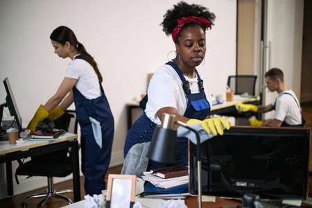 Black young adult woman cleaning computer monitor with cloth and gloves in modern office, while Caucasian young adult woman and Caucasian young adult man cleaning desks in backgroundの写真素材