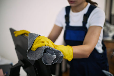 Young adult Caucasian woman wearing yellow gloves cleaning computer monitor with microfiber cloth in...