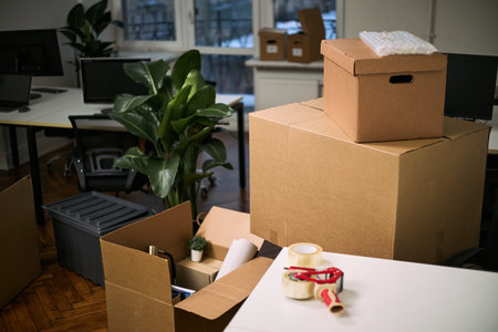 Stacked cardboard boxes and cleaning supplies occupying modern office workspace, packing materials...