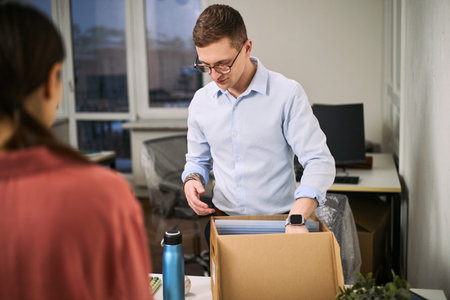 Caucasian young adult man packing office supplies into cardboard box while standing at desk, brown haired young adult woman assisting with cleaning and organizing workspaceの写真素材