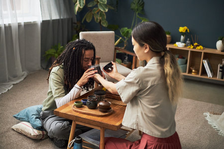 Young adult Asian woman performing traditional tea ceremony with young adult Black woman sitting on floor, both engaging in cultural ritual, tea set arranged on wooden tray