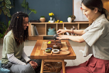 Asian young woman pouring tea for Black young man during traditional tea ceremony, both sitting at wooden table in modern living room, engaging in cultural exchange and conversation