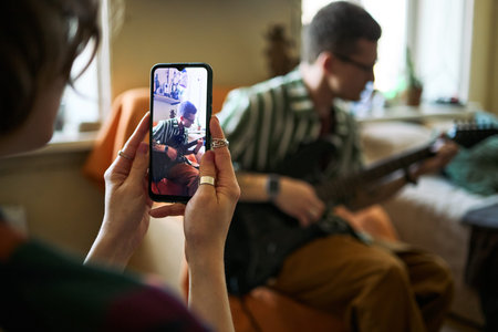 Young adult woman recording young adult man playing electric guitar on smartphone at home, creating social media music content. Useful for creator economy, mobile video marketing, close up