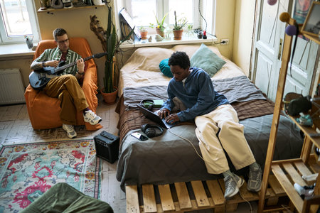 High angle view of young adult men collaborating at home, one playing electric guitar, one working on laptop, showing music production and creative remote workflow for content marketing