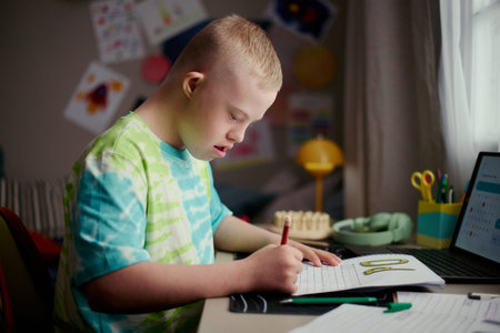 Caucasian boy with Down Syndrome writing workbook exercises at desk, following online lesson on laptop and practicing literacy skills. Useful for inclusive education, homeschooling, child development