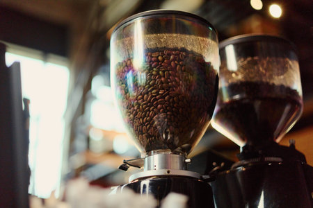 Close up shot of roasted coffee beans filling transparent hopper of professional electric grinder in cafe. Visual concept suitable for coffee shop branding and beverage advertising